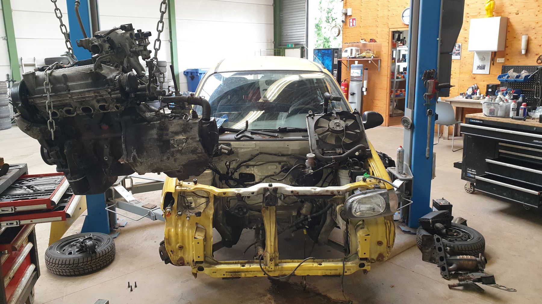 Een motor hangt aan de takel boven een gele auto zonder voorkant